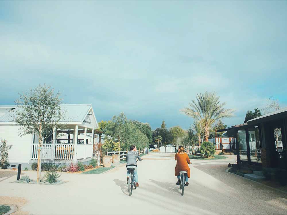 Two female cyclists leisurely pedal down a dirt road flanked by vintage buildings.