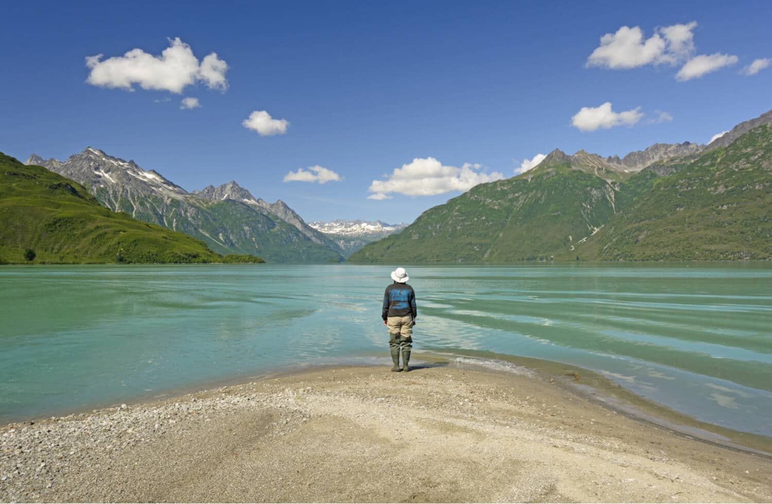 Camper with wading boots stares out on a lake surrounded by mountains.