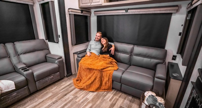 A young couple with laps covered by a blanket relax on a couch in an RV.