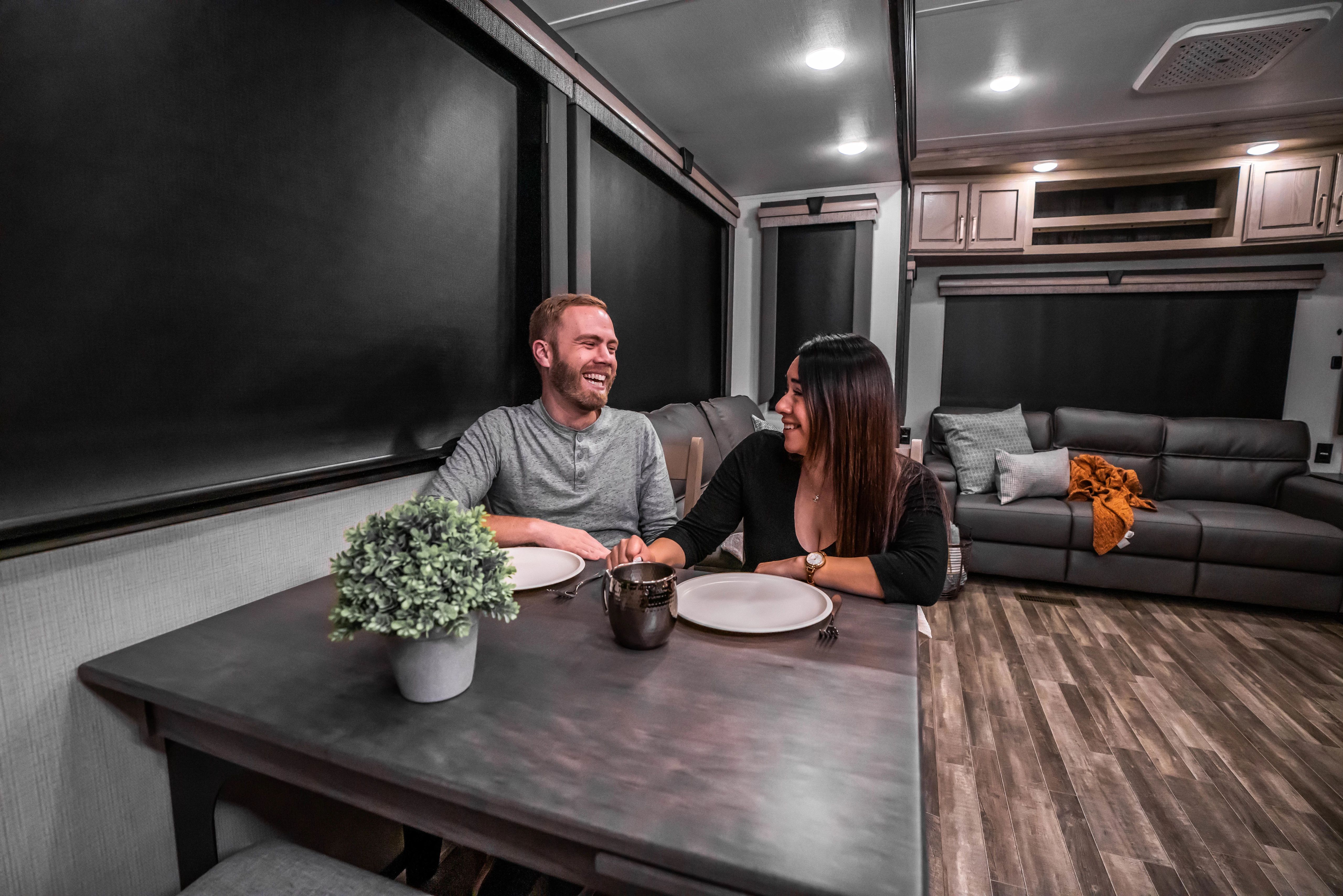 A young couple share a laugh seated at a dining room table in an RV.