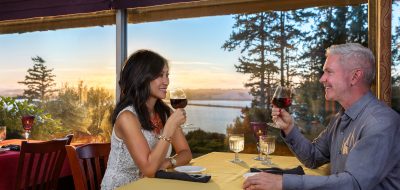 A couple enjoy a glass of wine in a restaurant with a window that looks out over a scenic coastal area.