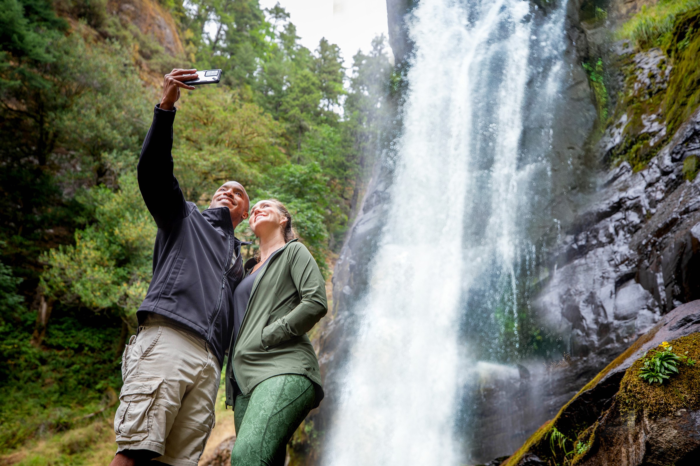 A couple pose for a selfie in front of a long waterfall.