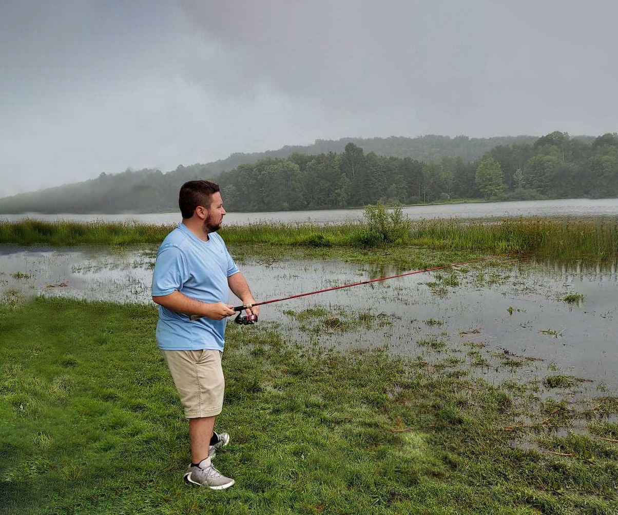 Man in shorts standing on a grassy bank with pole on an overcast day.