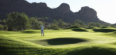 Woman swinging golf club in lush course fringed by rugged mountains.