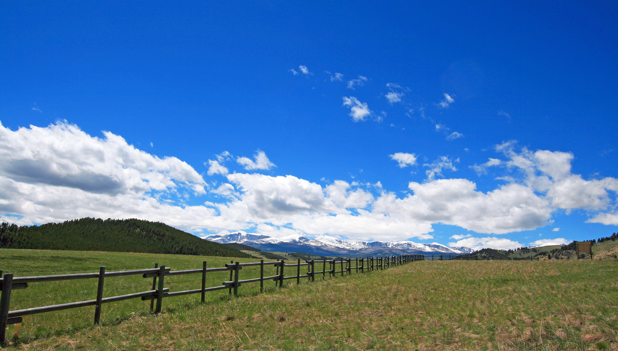 A wooden fence cuts across a sprawling field fringed by hills under a blue sky.