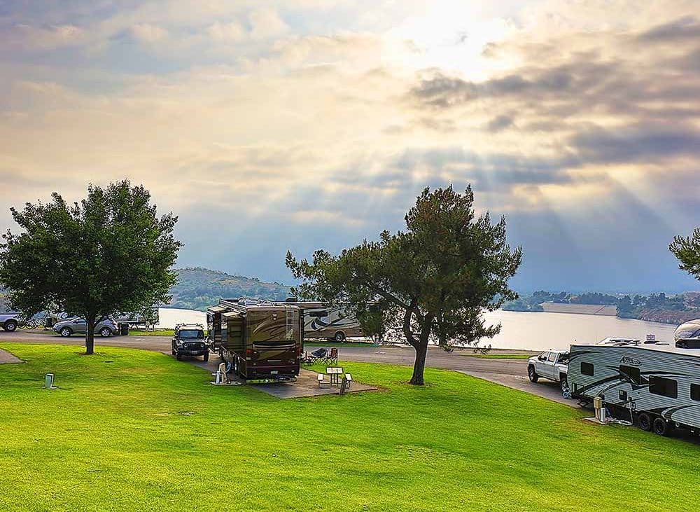 Sun breaks through the clouds and pours its rays onto a lake and a grassy RV resort on the banks.