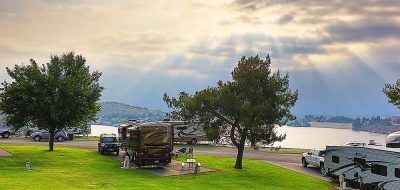 Sun breaks through the clouds and pours its rays onto a lake and a grassy RV resort on the banks.