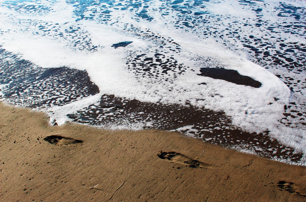 Footsteps imprinted on a seashore as water laps the sand.