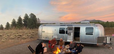 A group of RV travelers gathered around a campfire in front of an Airstream Motorhome.