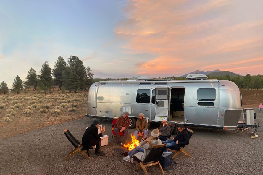 A group of RV travelers gathered around a campfire in front of an Airstream Motorhome.