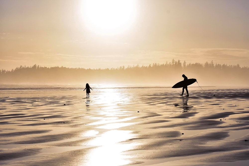 A surfer walks on shimmering sands.