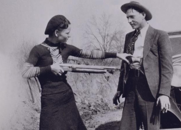 A black and white photo of a woman holding a shotgun to a man in a fedora.