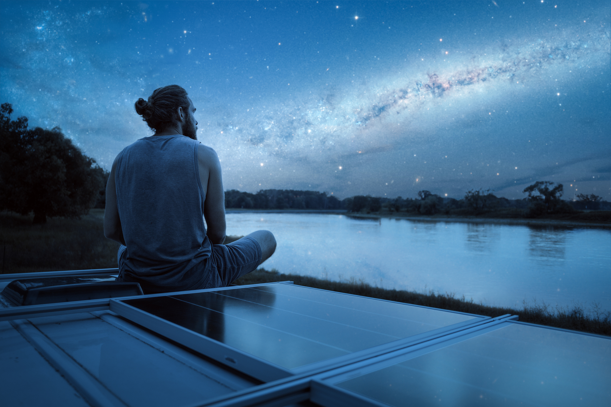 A young man is sitting on the roof of a camper van during nighttime with a beautiful starry night sky above him.