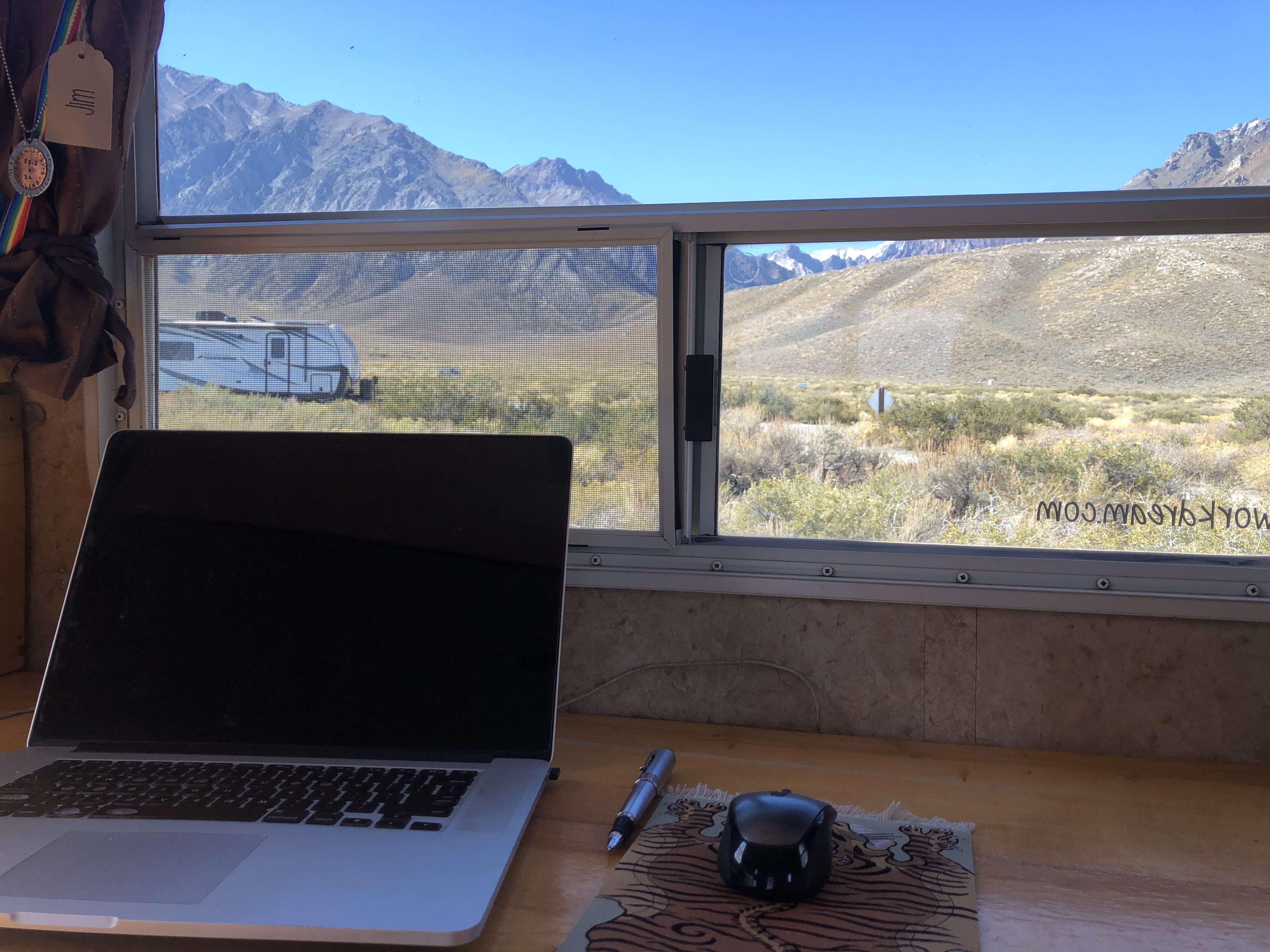 A laptop on a desk overlooking a landscape of rocky mountains, rolling hill, desert scrub and lone trailer.