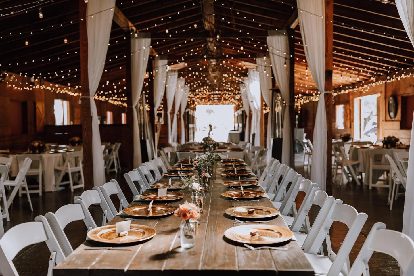 A long dining table flanked by white chairs in a barn setting.