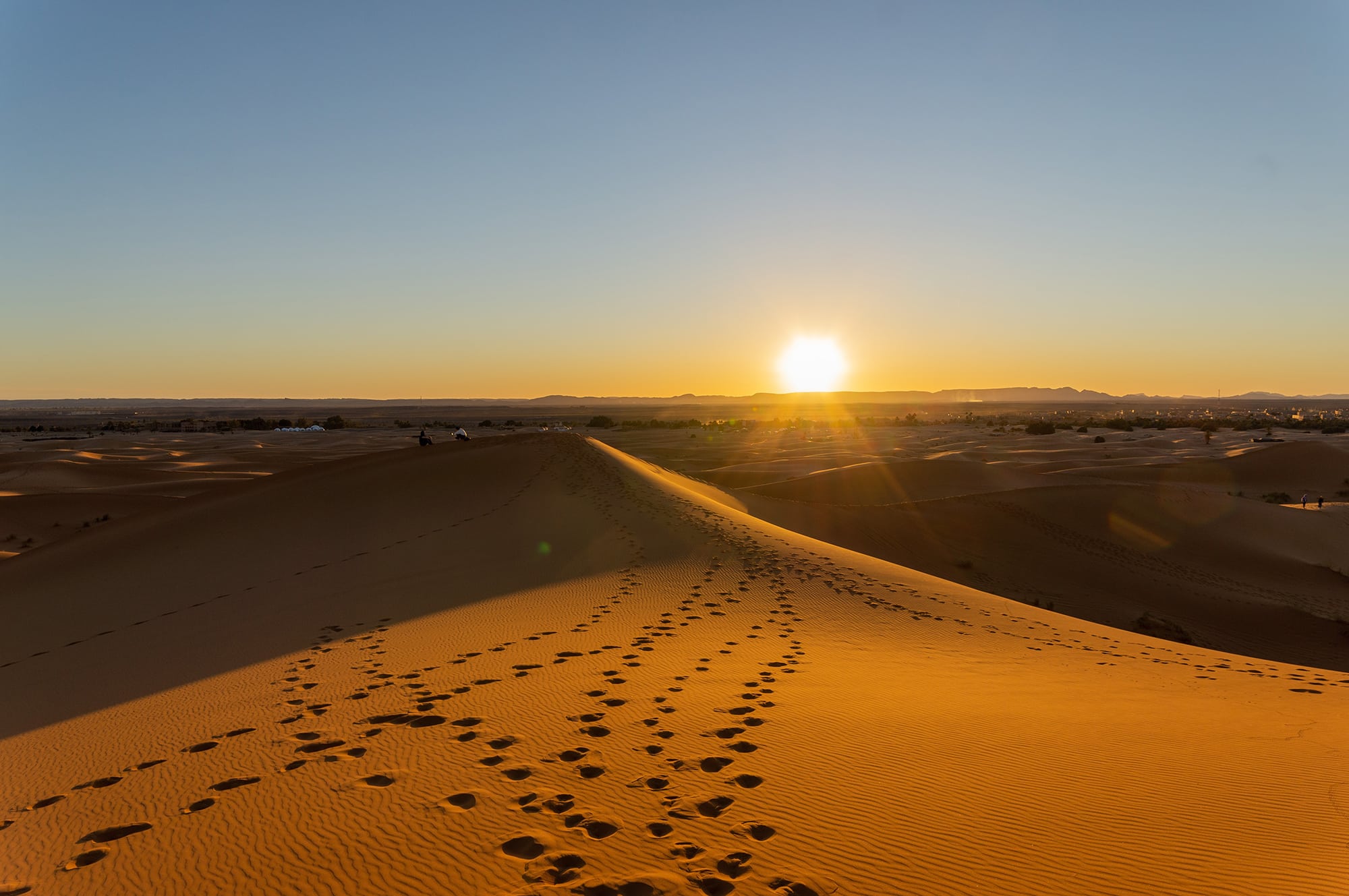 A trail of footsteps crossing a dune with sun on horizon.
