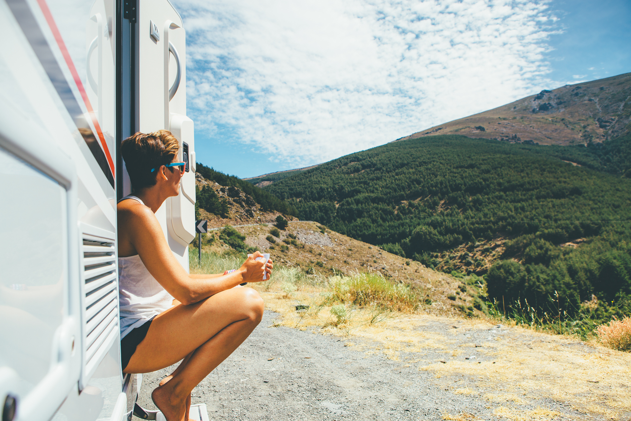 A vista lateral de uma jovem está sentada em um degrau de caravana e segurando uma xícara em uma parada de viagem de aventura de férias.  Copie a área de espaço disponível