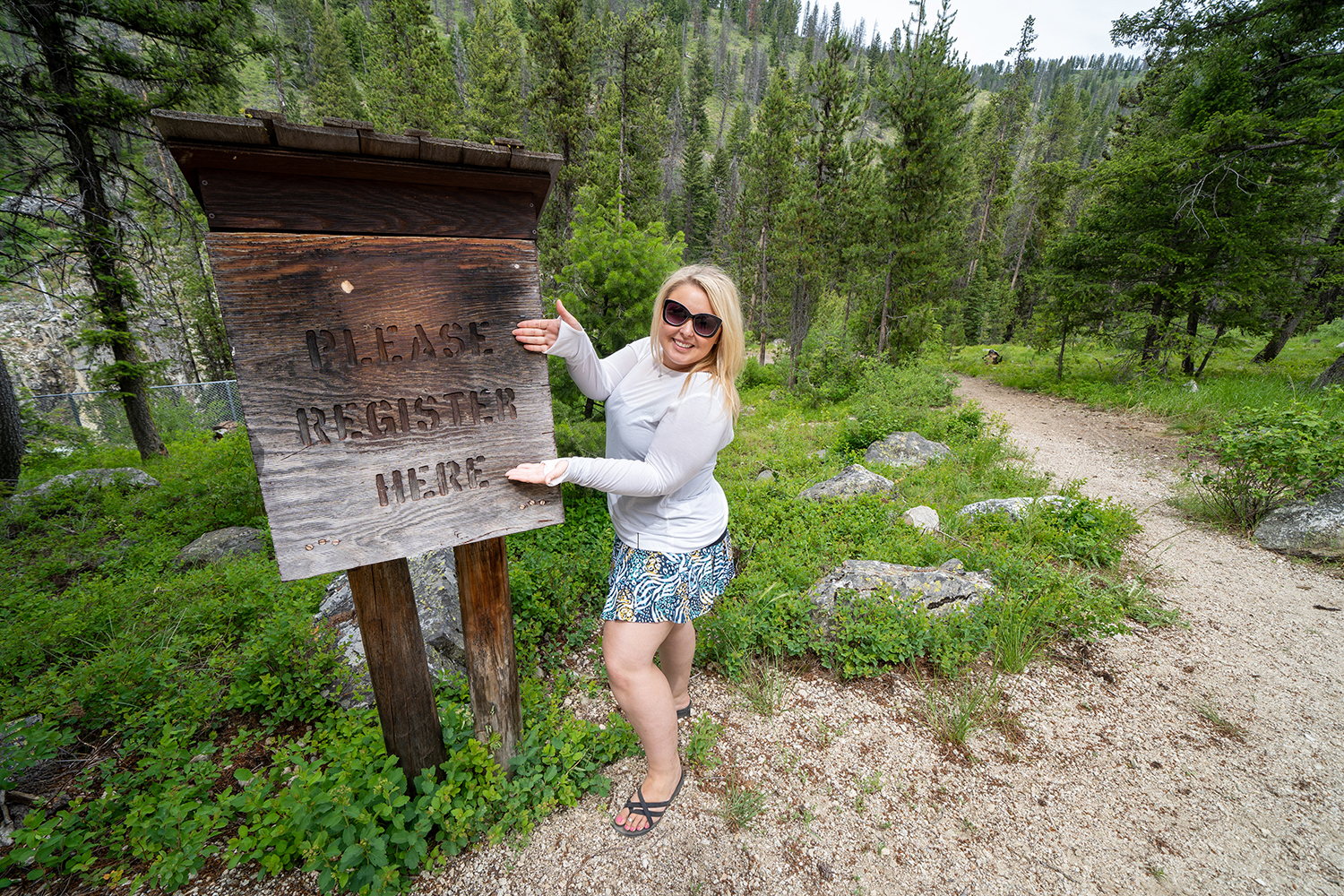 Blonde woman poses near a sign for campers and hikers to register - Please Register Here - at a campground in the Sawtooth Wilderness National Forest in Idaho