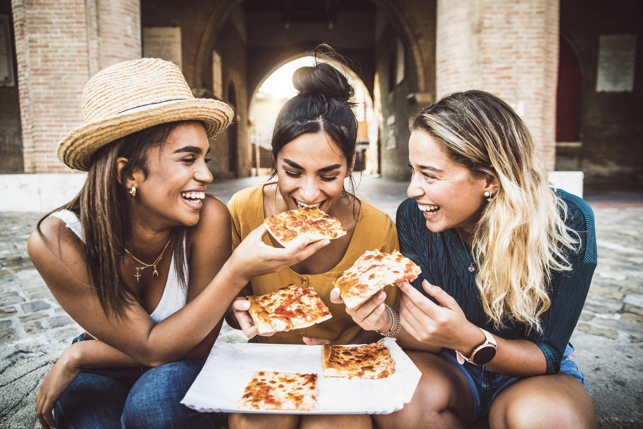Three young women enjoy pizza on a city street.