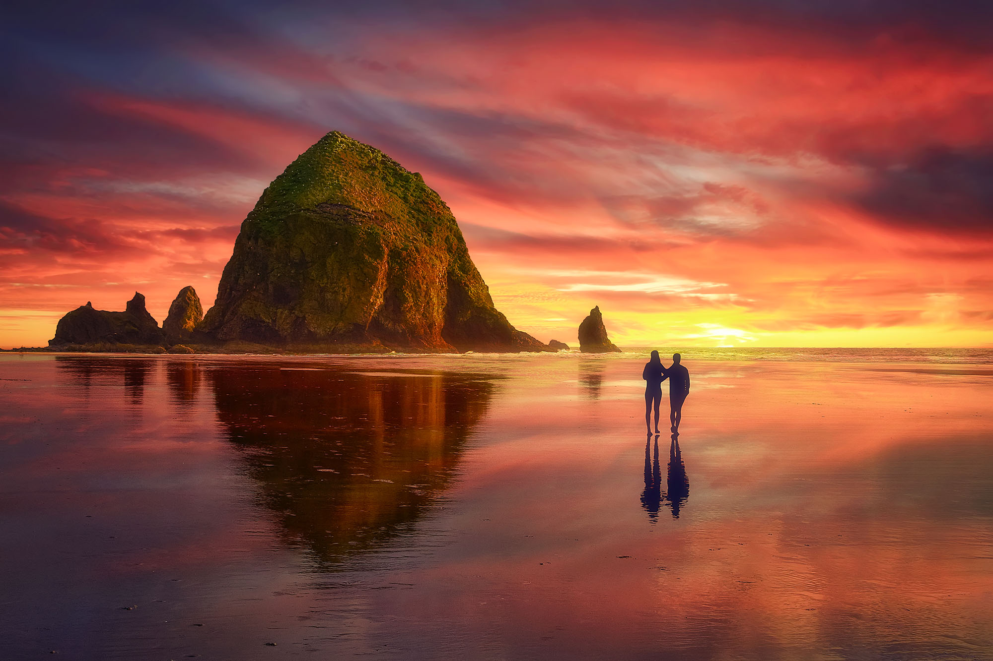 Rock outcropping looms over a lone couple strolling on a glassy sunset beach.