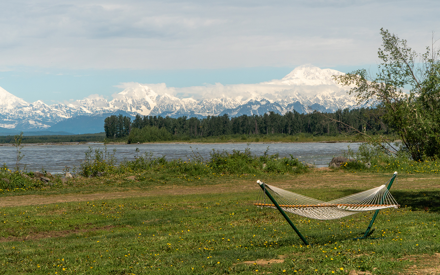 Hammock erected on riverbank with snow-capped mountains looming over horizon.
