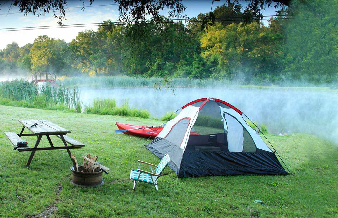 A dome tent on a grassy river bank as mist rises above the water.